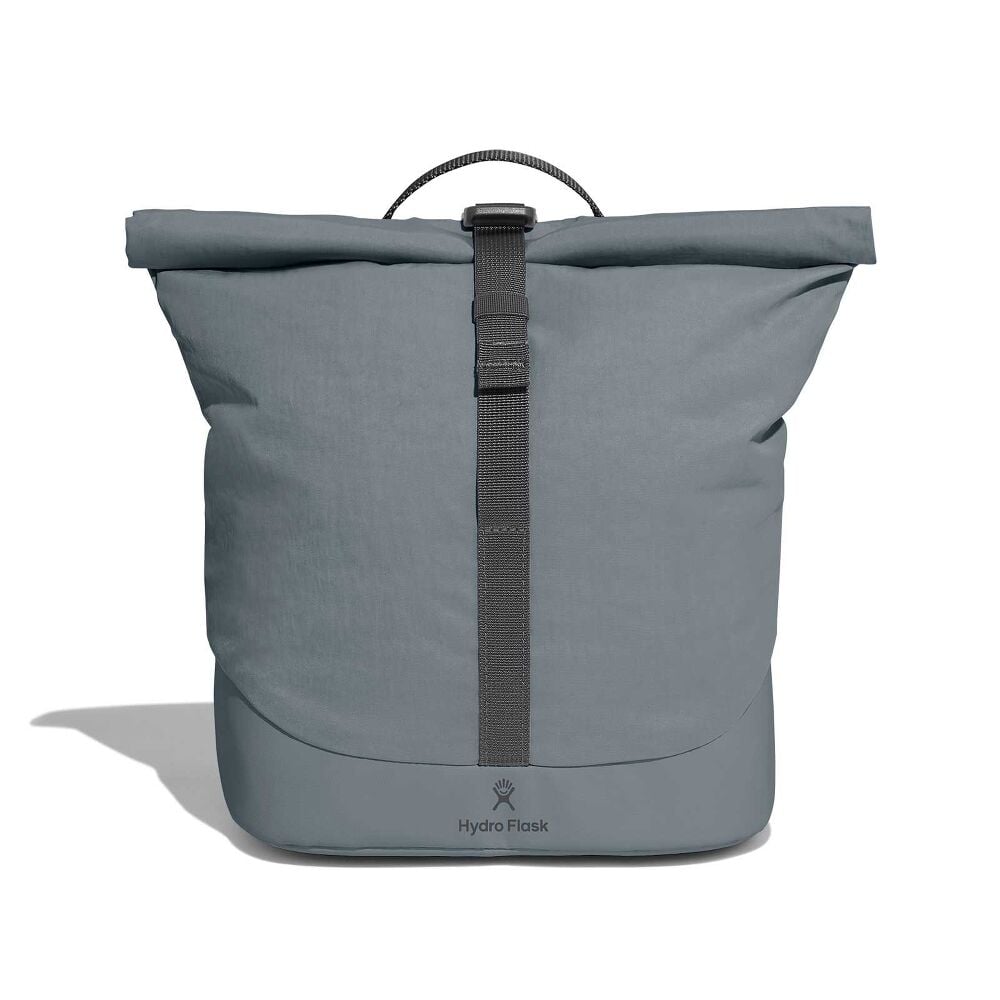 Person in olive sweater and dark jeans holding compact gray Hydro Flask roll-top bag with vertical black strap, buckle on front, and logo near the bottom.